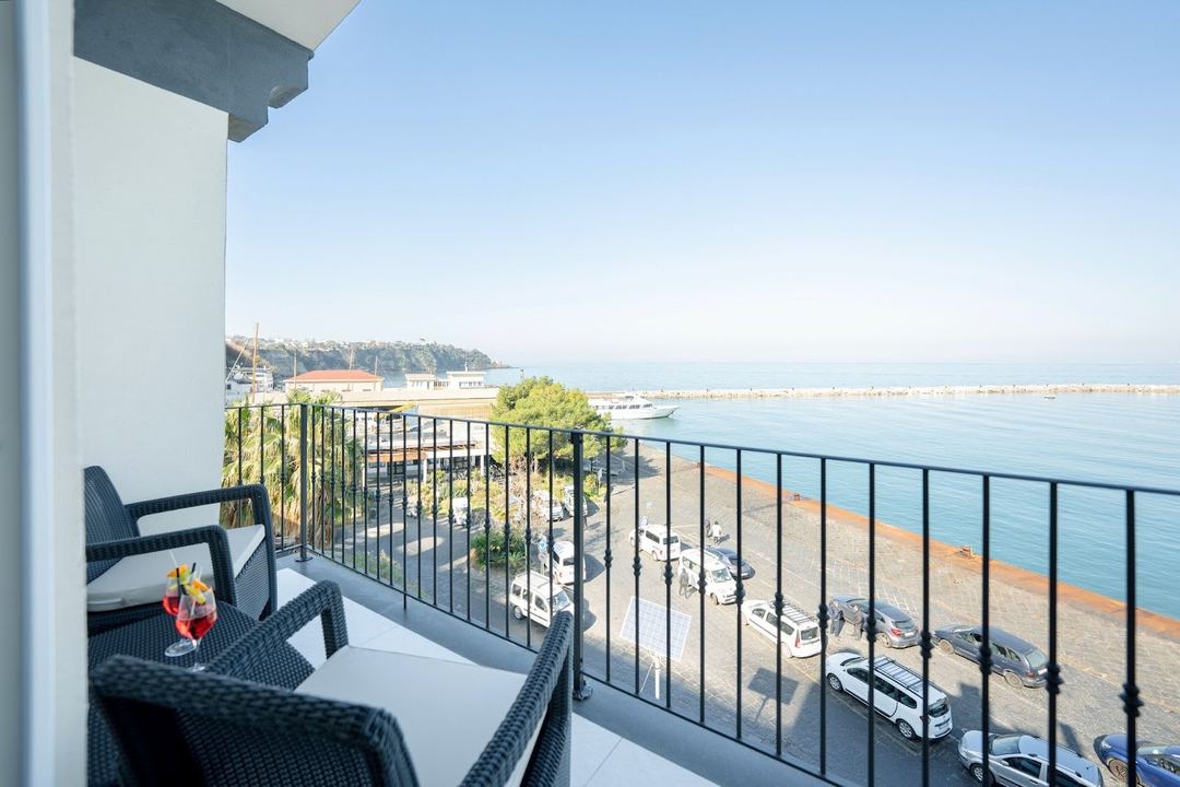 A balcony with comfortable seating overlooks a beach, marina, and sparkling Mediterranean Sea with distant coastline under clear skies.