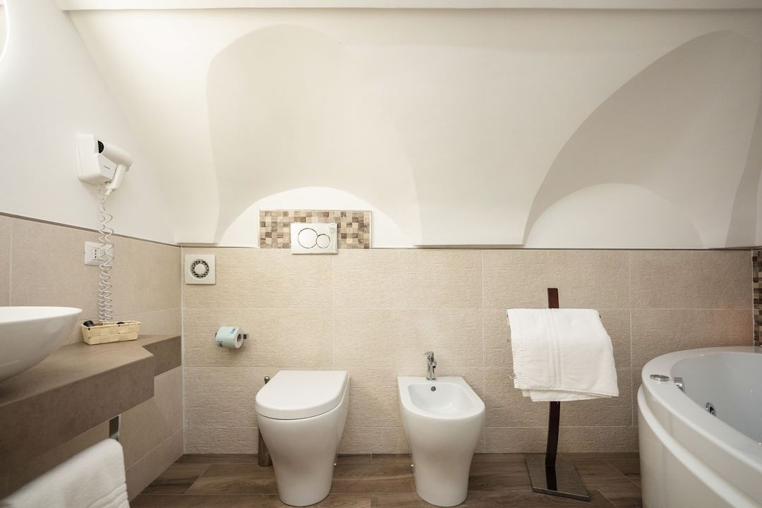 A modern bathroom with arched ceilings features a toilet, bidet, bathtub, and pedestal sink accented with neutral tile finishes and warm lighting.