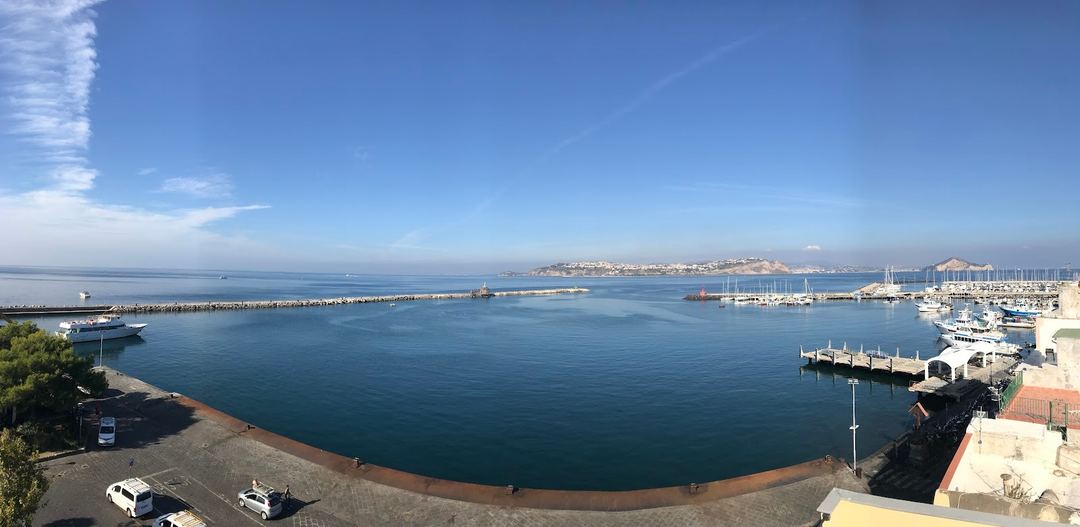 Panoramic view of Procida's harbor with sailboats, marina facilities, and the coastal town across the blue Mediterranean Sea under clear skies.